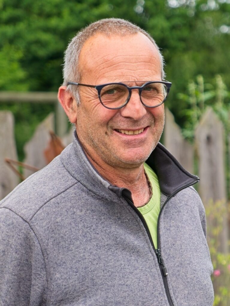 Portrait eines freundlich blickenden Mannes mit Brille und grauer Fleecejacke vor grünem Hintergrund im Außenbereich.