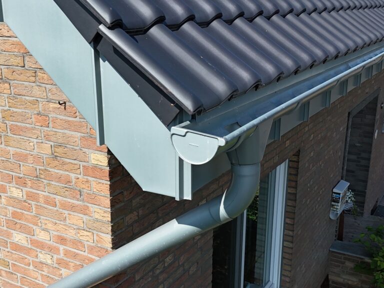 Detailaufnahme eines Ortgangs mit dunkelgrauen Ziegeln und vorbewitterter Rheinzink-Dachrinne samt Fallrohr an einer Klinkerfassade.